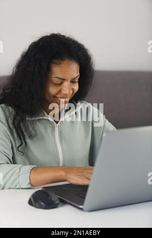 Happy African woman working on modern laptop computer at home. Cheerful BIPOC female typing text on modern notebook pc Stock Photo