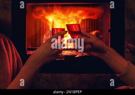Glasses of wine in hands of lovers on background of fire in fireplace. Coziness, love, celebration of Valentine's Day. Stock Photo