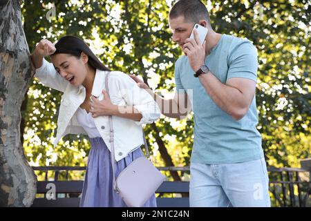 Man calling ambulance to help woman with heart attack in park Stock Photo