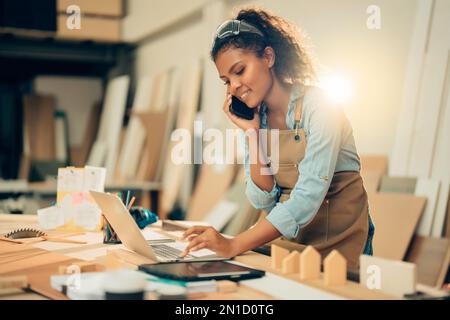 Smiling Young Asian female carpenter using laptop computer while talking to customer on the phone in woodcraft carpentry workshop. Stock Photo