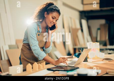 Smiling Young Asian female carpenter using laptop computer while talking to customer on the phone in woodcraft carpentry workshop. Stock Photo