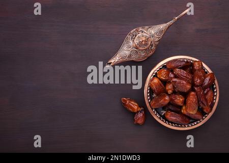 Plate with dates  and aladdin lamp on dark brown painted wooden background  with empty space for text . Flat lay. Ramadan background. Stock Photo