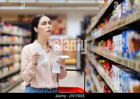 Portrait of young amazement Caucasian woman holds paper receipts of purchase and look at showcase of supermarket. In background, rows of shelves with Stock Photo