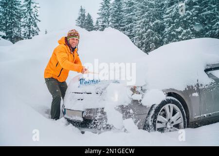 Smiling man in funny nepalese hat removing snow from car hood using big snow brush. The automobile freezes out on the countryside forest home driveway Stock Photo