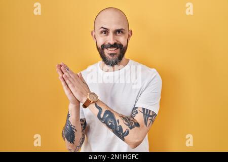 Young hispanic man with tattoos standing over yellow background clapping and applauding happy and joyful, smiling proud hands together Stock Photo