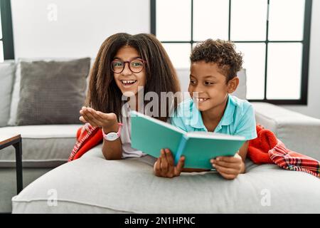 Two siblings lying on the sofa reading a book pointing aside with hands open palms showing copy space, presenting advertisement smiling excited happy Stock Photo