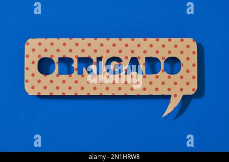 a paper sign in the shape of a speech bubble with the text thank you written in portuguese, on a blue background Stock Photo