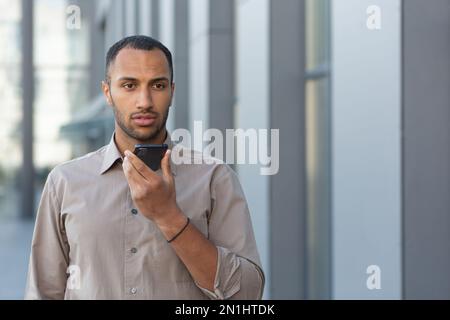 Angry businessman in shirt recording audio message from outside office building, african american man using assistant giving commands to artificial intelligence using smartphone. Stock Photo