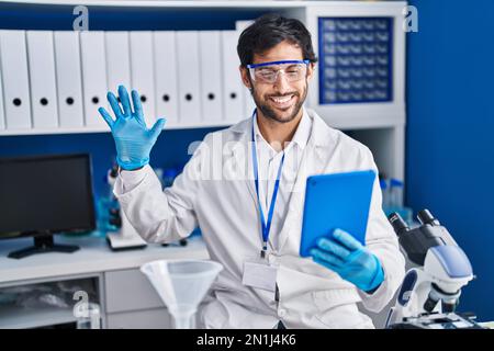 Handsome latin man working at scientist laboratory using tablet looking positive and happy standing and smiling with a confident smile showing teeth Stock Photo