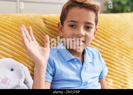 Adorable hispanic boy saying hello with hand sitting on sofa at home Stock Photo