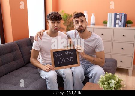 Homosexual gay couple holding blackboard with first home text puffing cheeks with funny face. mouth inflated with air, catching air. Stock Photo