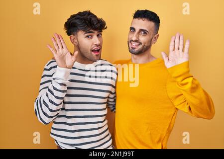 Young hispanic gay couple standing over yellow background waiving saying hello happy and smiling, friendly welcome gesture Stock Photo