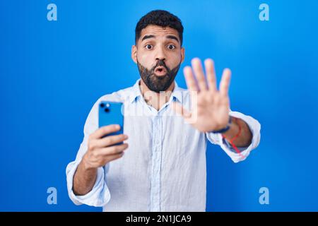 Hispanic man with beard using smartphone typing message doing stop gesture with hands palms, angry and frustration expression Stock Photo