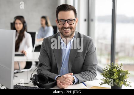 Smiling bearded businessman wearing eyeglass sitting in call center office Stock Photo