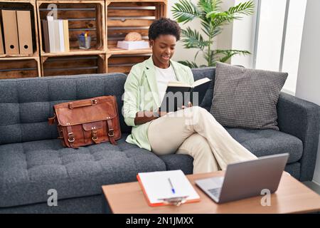 African american woman psychologist reading book sitting on sofa at psychology center Stock Photo
