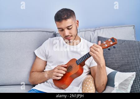 Young hispanic man playing ukelele sitting on sofa at home Stock Photo