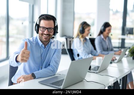 Handsome male online support agent with headset showing thumb up accompanied with his colleagues at call center. Stock Photo