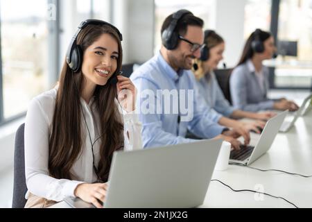 Beautiful friendly online support agent with headset working in row with her colleagues operators at customer service office. Stock Photo