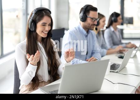 Smiling female call center operator in headphones with microphone consulting client on phone in customer support service while working with group of c Stock Photo