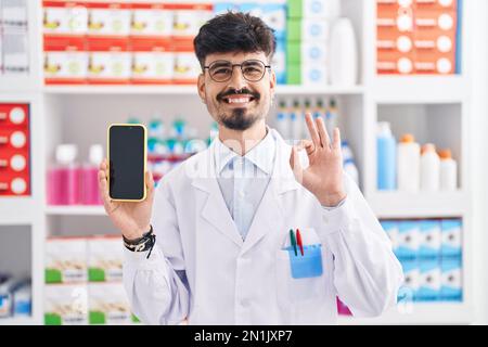 Young hispanic man with beard working at pharmacy drugstore showing smartphone screen doing ok sign with fingers, smiling friendly gesturing excellent Stock Photo