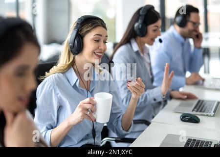 Smiling female customer support operator in headset talking to client accompanied with coworker operators at call center Stock Photo