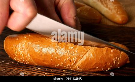Chefs hands slicing a hot dog bun with sesame seeds with kitchen knife in half. Preparation tasty fast food on domestic kitchen. Selective focus. Clos Stock Photo