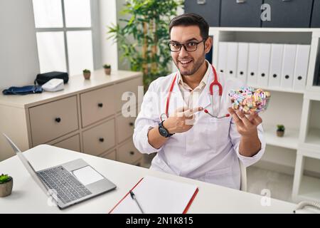 Young hispanic doctor man with beard holding candy smiling happy pointing with hand and finger Stock Photo