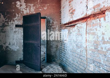 Old store-room storeroom in the bakery at Kolmanskop Ghost Town, Namibia.There's a rusted metal door and peeling wall paint. Creative, processed image. Stock Photo