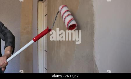 red and white roller on a long handle in the process of applying a layer of glue on a plastered wall in a home room, priming room walls Stock Photo