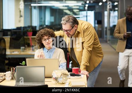 Young confident businesswoman in casualwear explaining something to mature male colleague while both looking at laptop screen Stock Photo