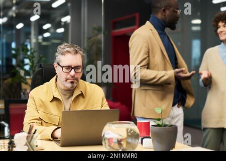 Serious mature businessman networking in front of laptop while sitting by desk against two young intercultural colleagues having discussion Stock Photo