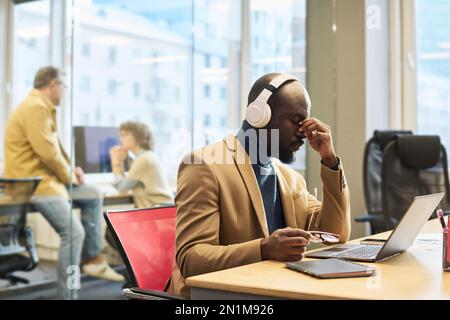 Young tired businessman in headphones sitting by workplace in front of laptop and having short break against his colleagues having talk Stock Photo
