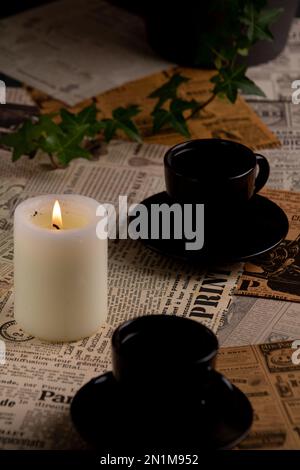photo two coffee cups with coffee standing on a background of old newspapers next to a burning candle Stock Photo