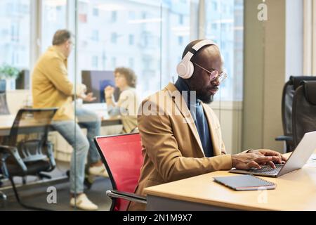 Serious African American employee with headphones sitting by desk in front of laptop and entering new data for business project Stock Photo