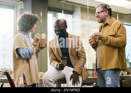 Happy young African American businessman with cup of coffee talking to colleagues eating Chinese wok at lunch break in openspace office Stock Photo