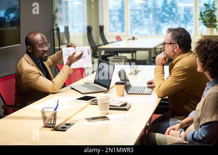 Confident African American broker presenting graphic financial data to his colleagues during discussion of marketing development Stock Photo