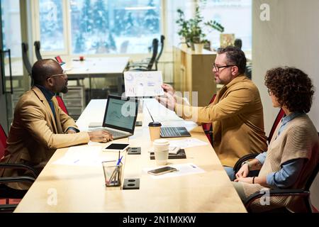Confident mature male analyst showing paper document with graphic financial data to young intercultural subordinates or colleagues Stock Photo