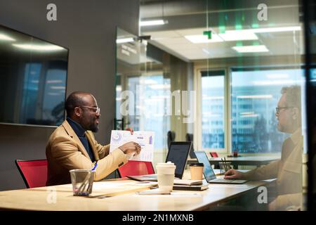 Confident male economist commenting graphic data on paper document while sitting by desk in front of colleague at working meeting Stock Photo