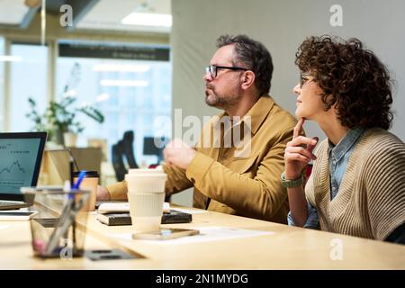 Two serious colleagues sitting by desk at seminar and listening to speaker making presentation of financial data or marketing analysis Stock Photo
