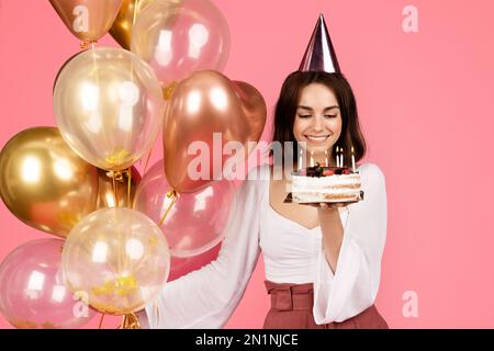 Happy pretty young european woman in hat holding many balloons and cake with candles, makes a wish Stock Photo