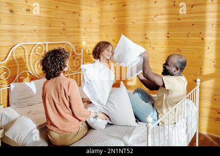 Happy young African American man with pillow fighting with adorable little son while having fun on bed after sleep in the morning Stock Photo