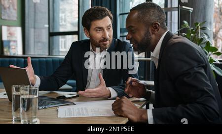 Two men amazed diverse workers with laptop in office talking emotional discuss project startup business result sitting at desk. Multiracial partners Stock Photo