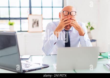 Middle aged businessman holding his head and pondering while sitting at the office and using laptop for work. Stock Photo