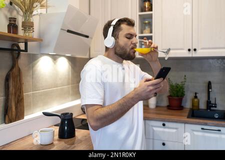 Male athlete in headphones drinking orange juice in morning and reading news on internet using his mobile phone. Stock Photo