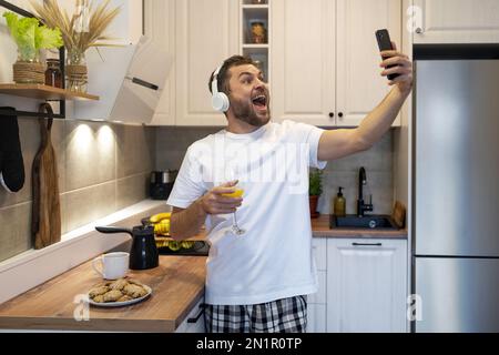 Man in headphones standing in kitchen in morning taking selfie using his phone and having fun. Stock Photo