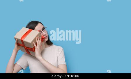 Portrait of a student girl with glasses and a beige blouse holding a large gift box with a red bow, trying to guess what's inside on a blue background Stock Photo
