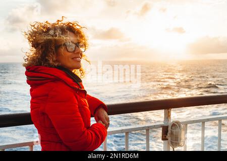 Cruise ship vacation woman enjoying sunset on travel at sea. Traveler happy woman in red jacket looking at ocean relaxing on luxury cruise liner boat. Stock Photo