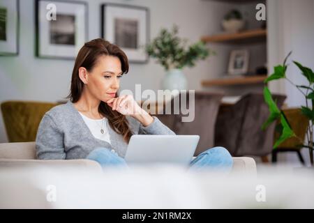 Shot of an attractive middle aged woman sitting in an armchair while using laptop at home. Blank screen. Beautiful female looking thoughtfully and wea Stock Photo