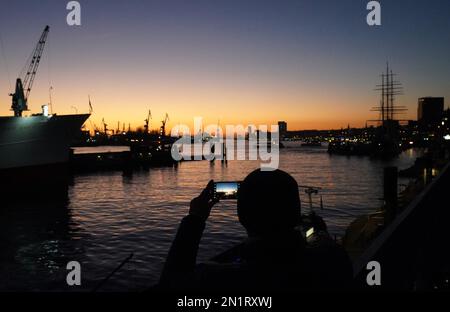 Hamburg, Germany. 06th Feb, 2023. A man photographs the evening atmosphere during sunset on the Jan Fedder promenade in the harbor. Credit: Marcus Brandt/dpa/Alamy Live News Stock Photo