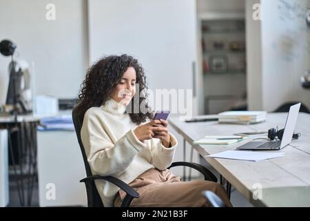 Smiling hispanic business woman using phone working in office sitting at desk. Stock Photo
